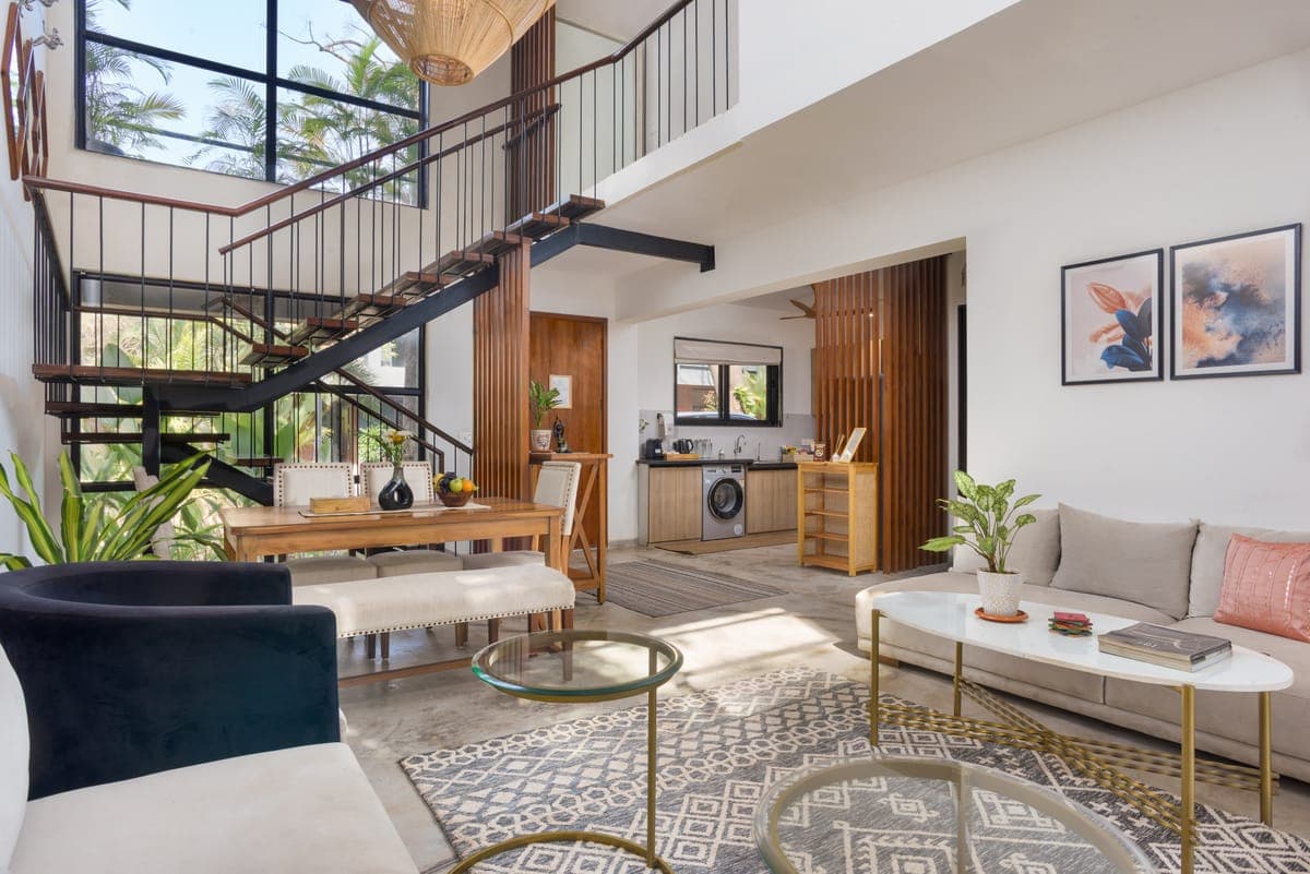 A double-height Nistula villa living room with a steel staircase, dining table and tall windows onto the palms.